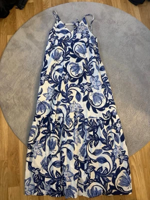 Blåvit långklänning med blommönster - Säljer en somrig långklänning med tunna axelband och v-ringning. Klänningen är vit med stora blå blommor och blad i ett härligt mönster.  Öppen rygg. Materialet känns lätt och luftigt, perfekt för varma dagar. Passformen är loose och klänningen har en avslappnad siluett. Jätte skön och super fin! Inte fått användning för!