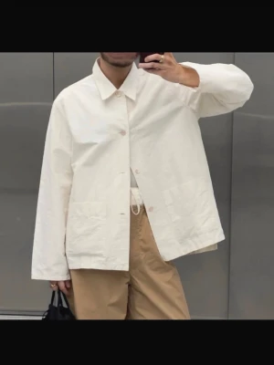 Vit overshirt jacka från Our Legacy - Stilren vit overshirt från Our Legacy i ett lätt, strukturerat bomullstyg. Rak passform med klassisk krage, knäppning framtill och två stora fickor. Perfekt för lager-på-lager och enkel att matcha med olika outfits. Enda defekten: viss missfärgning på kragen.