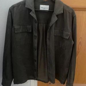 Olivgrön overshirt från These Glory Days - Snygg olivgrön overshirt från These Glory Days med klassisk krage och två stora bröstfickor med lock. Jackan har knäppning framtill och är tillverkad i ett mjukt bomullstyg. Perfekt för lager-på-lager och enkel att styla till olika outfits. Storlek m men sitter som s