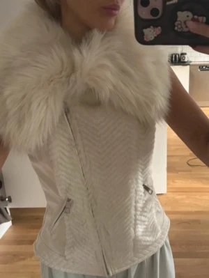 Vit pälsväst från Guess - Snygg vit pälsväst från Guess med stor fluffig fuskpälskrage och dragkedja framtill. Västen har en figurnära passform och två fickor med dragkedja. Perfekt för att lyfta din outfit med en lyxig touch.