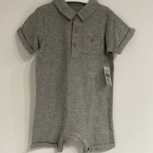 Grå kortärmad jumpsuit med krage 12-18 months baby - Snygg grå jumpsuit från Bebby med korta ärmar, krage och träknappar framtill. Har en bröstficka och uppvikta ärmslut. Tillverkad i mjukt, stretchigt material som känns skönt mot huden. Perfekt för en avslappnad och cool look.