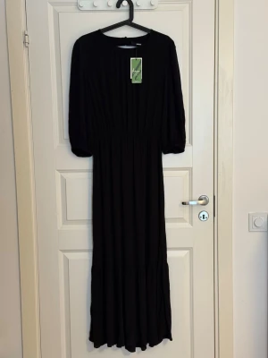 Svart långklänning från bikbok - Svart långklänning från bikbok med rund halsringning och trekvartsärmar. Djup urringning med knytband baktill. Klänningen har en markerad midja och ett mjukt, flytande fall. Tillverkad i ett lätt och bekvämt material som passar perfekt för en stilren look.