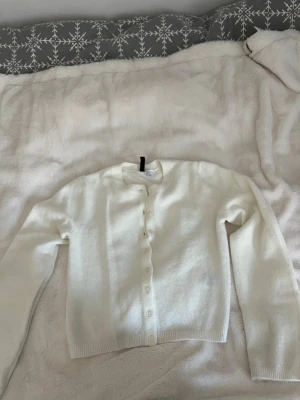 Vit kofta från Divided H&M - Säljer en enkel och stilren vit kofta från Divided H&M. Koftan har rund halsringning, knappar framtill och är ribbstickad vid ärmslut och nederkant. Perfekt att slänga på sig över en topp för en clean look. Koftan är något kortare i modellen. På ena armen är det en liten fläck (se bild)