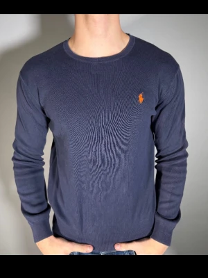 Mörkblå tröja från Ralph Lauren - Mörkblå långärmad tröja från Ralph Lauren med rund halsringning och den klassiska orangea logotypen broderad på bröstet. Tröjan är tunnstickad och har ribbade muddar vid ärmslut och nederkant. Perfekt för en clean och stilren look.