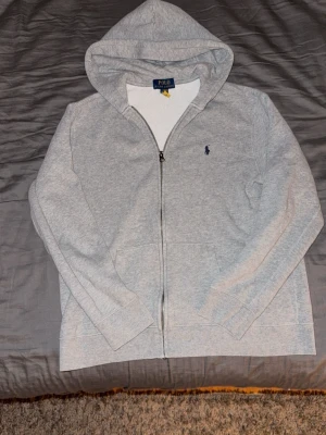 Grå zip hoodie från Polo Ralph Lauren - Snygg grå hoodie med dragkedja från Polo Ralph Lauren. Klassisk design med huva, fickor framtill och den ikoniska lilla blå loggan på bröstet. Tillverkad i mjukt sweatshirtmaterial som är perfekt för chill dagar. Enkel att matcha med jeans eller joggers.