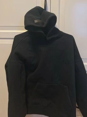 Svart Essentials hoodie med huva - Säljer en svart hoodie från Essentials med stor huva och känguruficka framtill. Diskret Essentials-logga på bröstet och större logga på ryggen. Tillverkad i mjukt bomullsmaterial, perfekt för en avslappnad och trendig look. Köpt på merchsweden, kvitto kan vidarebefodras vid köp. Storlek S men passar även M.