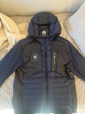 Mörkblå pufferjacka Sail Racing - Snygg mörkblå pufferjacka från Sail Racing med huva och flera logopatchar på bröstet och ärmen. Jackan har quiltad design, dragkedja framtill och är perfekt för kalla vinterdagar. Materialet är glansigt och ger en sportig look.