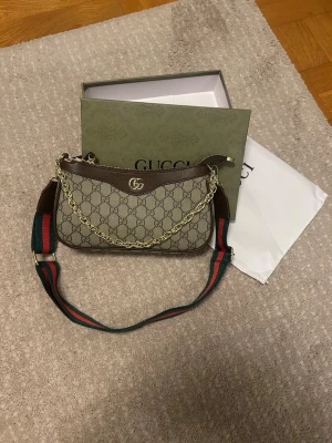 Gucci GG Supreme axelremsväska - Säljer en snygg Gucci GG Supreme handväska i beige och brunt med klassiskt GG-mönster. Väskan har guldfärgad kedjedetalj, bred axelrem i grönt och rött samt detaljer i brunt läder. Perfekt för dig som vill ha en ikonisk och trendig accessoar.