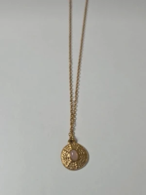 Guldigt halsband i rostfritt stål med rund berlock - Snyggt halsband i guldfärgat rostfritt stål med en rund, hamrad berlock. Berlocken har en ljus pärlliknande detalj i mitten som ger en elegant touch. Kedjan är tunn och justerbar för perfekt passform. Perfekt accessoar för att lyfta din outfit.