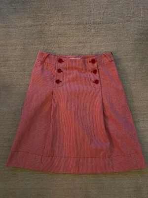 Strl 36 Röd- och vitrandig kjol med knappar - Snygg kjol  i röd- och vitrandigt tyg med sex dekorativa knappar framtill. Kjolen har en lätt A-linjeform och är tillverkad i ett mjukt bomullsmaterial. Perfekt för dig som gillar en retroinspirerad stil.