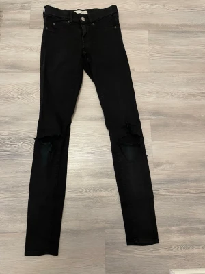 Svarta skinny jeans med hål - Säljer ett par svarta skinny jeans från Gina Tricot med slitna hål på båda knäna. Jeansen har klassisk femficksmodell, hög midja och smal passform. Materialet är stretchigt för extra komfort och de har knapp- och dragkedjestängning. 