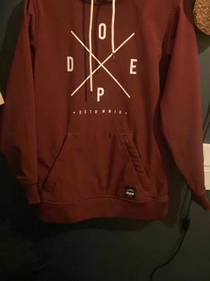 Vinröd hoodie från Dope, XS - Säljer en vinröd hoodie från Dope med vit grafisk print på bröstet och vita dragsnören i huvan. Hoodien har en stor magficka och ribbade muddar. Mjuk insida för extra komfort. Perfekt för dig som gillar streetwear och snygga detaljer.
