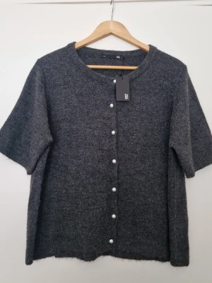 Mörkgrå kofta med pärlknappar från  Lager 157 - Kortärmad mörkgrå kofta från Lager 157 med rund halsringning och dekorativa pärlknappar framtill. Koftan är stickad i ett mjukt material och har en avslappnad passform som ger en chill vibe. Perfekt att slänga över en topp för en enkel men snygg look. Storlek L. Ny med prislapp.
