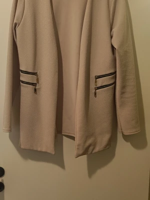Beige kavaj med dragkedjedetaljer - Stilren beige kavaj med öppen front och fyra dekorativa dragkedjor vid fickorna. Jackan har lång ärm och rak passform, tillverkad i ett mjukt och strukturerat material som ger en clean look. Perfekt för dig som gillar minimalistisk stil och snygga detaljer.