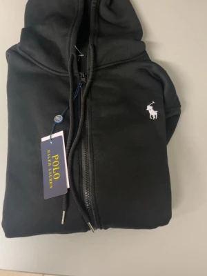 Svart kofta från Polo Ralph Lauren - Snygg svart kofta med dragkedja från Polo Ralph Lauren. Klassisk design med vit broderad logga på bröstet, huva med dragsko och två fickor framtill. Tillverkad i mjukt material som känns skönt mot huden. Perfekt för en avslappnad och stilren look.