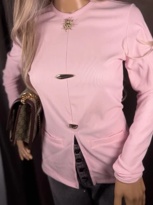 Ljusrosa ribbad långärmad topp - Trendig ljusrosa ribbad topp med lång ärm och unika dekorativa metallknappar framtill. Toppen har rund halsringning och två fickor framtill. Snygg detalj med broderad brosch vid halsen och slits nedtill.