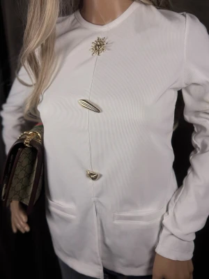 Vit blus med unika gulddetaljer - Stilren vit blus med lång ärm och rund halsringning. Blusen har dekorativa guldfärgade knappar och en broderad brosch framtill som ger en exklusiv känsla. Materialet är tunt och lätt ribbat för en modern look. Perfekt för dig som gillar detaljer och vill sticka ut.