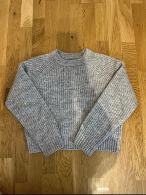 Grå stickad tröja från Lindex - Mysig grå stickad tröja från Lindex med ribbstickad struktur och rund hals. Tröjan har långa ärmar och breda muddar vid ärmslut och nederkant. Perfekt för kalla dagar och enkel att matcha med jeans eller kjol.