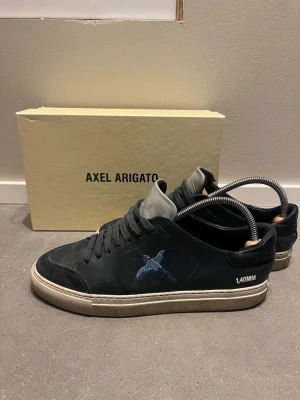 Marinblåa Axel Arigato skor  - Marinblåa sneakers från Axel Arigato med blått broderat fågelmotiv på sidan och vit sula. Skorna har mörkblåa skosnören, grå plös och detaljer i mocka och läder. Snygg och stilren design med texten '1.40MM' på sidan. Skorna har ett litet hål ovanför hälkappan men sitter fint ändå. Pris kan diskuteras vid intresse av skorna. 