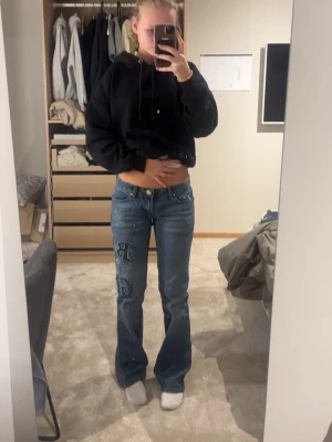 Blå bootcut jeans - Säljer ett par blå bootcut jeans med broderade detaljer på vänster ben. Jeansen har låg midja, klassiska fickor och en snygg vintagekänsla. Perfekta för dig som gillar en avslappnad men trendig stil. Midjemått: ca 40cm, innerbenslängd: ca 82cm