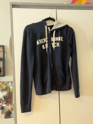 Marinblå hoodie Abercrombie & Fitch - Snygg marinblå hoodie från Abercrombie & Fitch med dragkedja och vita snören i huvan. Stor logga i vitt framtill och två fickor. Insidan av huvan är ljus och materialet känns mjukt och bekvämt, perfekt för chill dagar.