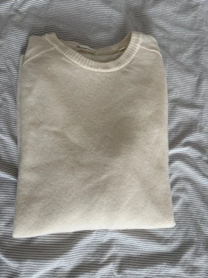 Beige stickad tröja från Arket - En stilren beige stickad tröja från Arket med rund halsringning och ribbade muddar vid ärmslut och nederkant. Tröjan har en mjuk och tunn stickning, perfekt för lager på lager och enkel att matcha med olika outfits.