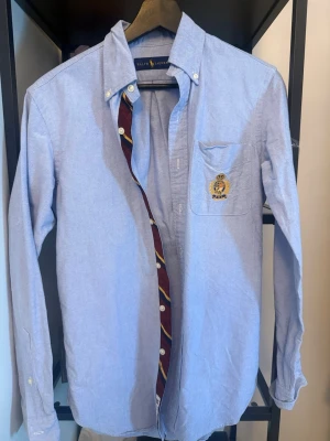 Blå skjorta från Ralph Lauren - Klassisk ljusblå skjorta från Ralph Lauren med broderad logga på bröstfickan. Skjortan har knappar hela vägen, button-down krage och lång ärm. Tillverkad i bomull och har en snygg detalj med randigt tyg på insidan av knappslån.