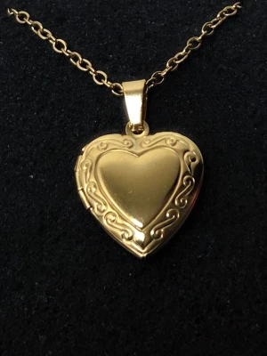 Guldfärgat hjärthalsband med medaljong Rostfritt Stål  - Snyggt halsband i guldfärg med ett hjärtformat hängsmycke som går att öppna som medaljong. Hängsmycket har fina snirkliga detaljer runt hjärtat och kedjan är av klassisk modell. Perfekt för att lägga in ett litet foto eller minne.