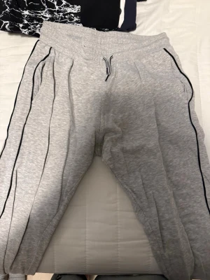 Grå mjukisbyxor med svarta detaljer - Säljer ett par grå mjukisbyxor med svarta ränder längs benen och dragsko i midjan. Byxorna har resår i midjan och vid bensluten samt praktiska sidofickor. Materialet är en mjuk bomullsblandning som är skön att ha på sig.