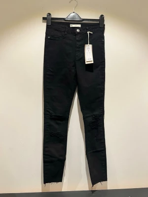 Jeans  - Säljer ett par svarta Molly jeans från Gina Tricot med slitningar över knäna och råa benslut. Jeansen har klassisk femficksmodell. Perfekta för dig som gillar en edgy och trendig stil.