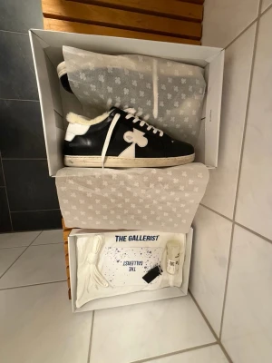 The Gallerist - Snygga svarta sneakers från The Gallerist med vit sula, vita skosnören och en vit klöverdetalj på sidan. Insidan är fodrad med mjuk fuskpäls för extra komfort. Perfekta för dig som vill ha en unik och trendig look. Fick de i julklapp men min farsa beställde fel storlek.