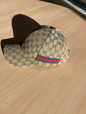 Beige GG keps från Gucci - Snygg beige keps från Gucci med klassiskt GG-monogram över hela kepsen. På sidan finns den ikoniska gröna och röda randen. Kepsen har böjd skärm och är tillverkad i canvasmaterial. Perfekt för dig som vill ha en exklusiv och trendig accessoar.