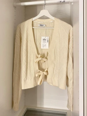 Kofta med knyt från ONLY - Säljer en krämvit stickad kofta från ONLY med snygg kabelstickad design och två knytband framtill. Koftan har djup V-ringning, ribbade muddar och en croppad passform. Perfekt för att lyfta din outfit med en söt och trendig touch.