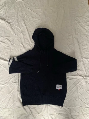 Mörkblå hoodie från Thom Browne - Snygg mörkblå hoodie från Thom Browne med klassisk huva och dragsko. Stickad struktur och ribbade muddar. Detaljerad med vita ränder på ena ärmen och en ikonisk etikett med fransk flaggdetalj på magen. Perfekt för en chill och stilren look.