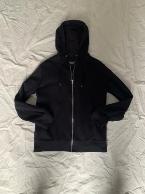 Svart hoodie med dragkedja från Primark - En svart hoodie från Primark med dragkedja framtill och justerbar huva med snören. Tillverkad i mjukt material som känns skönt mot huden. Perfekt för chill dagar eller när du vill ha en enkel och clean look.