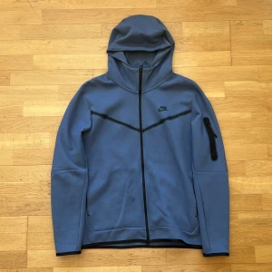Blå zip hoodie från Nike - Snygg blå hoodie från Nike med dragkedja framtill och svart Nike-logga på bröstet. Hoodien har huva, svarta detaljer längs bröstet och en praktisk ficka med dragkedja på ena ärmen. Perfekt för en sportig och avslappnad stil.
