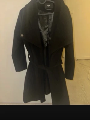 Svart kappa med skärp från Bik Bok - Stilren svart kappa från Bik Bok med bred krage och knytband i midjan. Kappan har slät yta, helsvart foder och en klassisk, rak passform. Perfekt för dig som vill ha en enkel men snygg look till hösten eller vintern.