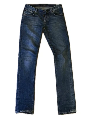Grim Tim jeans från Nudie Jeans - Snygga blå jeans från Nudie Jeans med slim ben och klassisk femficksmodell. Jeansen har slitningar och kontrastsömmar samt detaljerade bakfickor med Nudie-loggan. Tillverkade i denim med en cool tvättad look. Litet hål baken. 