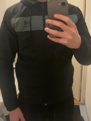 Svart hoodie från Under Armour - Snygg svart jacka från Under Armour med dragkedja framtill och en grå panel över bröstet där loggan syns. Hoodien har huva med dragsko och är tillverkad i ett mjukt material som passar perfekt till chill eller träning.