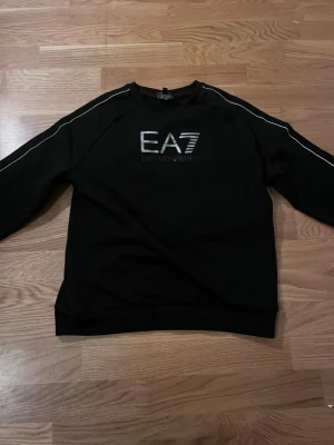 Svart sweatshirt från EA7 Armani - Snygg svart sweatshirt från EA7 Emporio Armani med tryckt logga på bröstet och vita detaljer längs ärmarna. Klassisk rund halsringning och mjukt material som passar perfekt till en avslappnad stil.