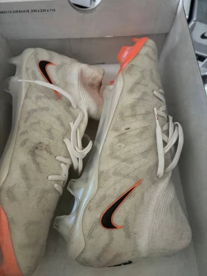 Nike vita fotbollsskor med orange detaljer - Säljer ett par vita Nike fotbollsskor med orange detaljer och svart Swoosh-logga. Skorna har en strömlinjeformad design, snörning och en lätt ovandel i syntetmaterial. Sulan har dobbar för bra grepp på planen. Perfekta för dig som vill sticka ut på matchen!