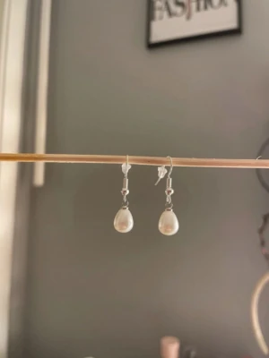 Silvriga örhängen med droppformade pärlor - Säljer ett par stilrena örhängen med silvriga krokar och droppformade vita pärlor. Enkla men ändå eleganta, perfekta för att lyfta din look. Passar dig som gillar klassiska accessoarer med en modern twist. Egengjorda
