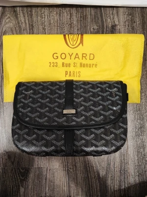 Svart axelremsväska  - Snygg svart kuvertväska med klassiskt vitt monogrammönster och detaljer i svart läder. Väskan har en elegant rektangulär form och stängs med ett lock och magnet. Perfekt för dig som vill ha en ikonisk accessoar med Paris-vibe.