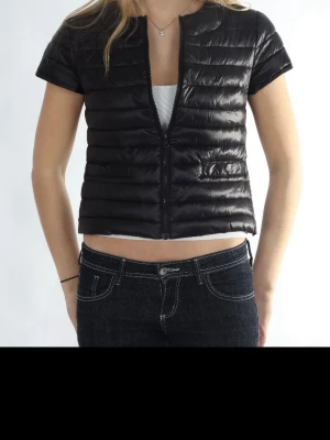 Kort svart dunväst med dragkedja - Snygg kort svart dunväst med horisontella sömmar och dragkedja framtill. Västen har korta ärmar och en croppad passform som ger en modern look. Tillverkad i glansigt material som ger en cool vibe. Perfekt att styla över en t-shirt eller hoodie.