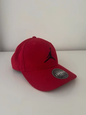 Röd Jordan keps med svart logga - Snygg röd keps från Jordan med den klassiska svarta Jumpman-loggan broderad framtill. Kepsen har böjd skärm och justerbar baksida. Tillverkad i slitstark bomull som ger en bekväm passform och sportig look.