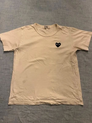 Beige t-shirt Comme des Garçons Play - Snygg beige t-shirt från Comme des Garçons Play med det ikoniska svarta hjärta-trycket på bröstet. Klassisk passform med rund hals och korta ärmar. Perfekt basplagg för en clean och stilren look. T shirten har 2 fläckar 