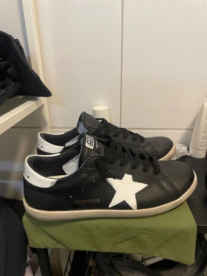 Svarta Golden Goose sneakers  - Svarta sneakers från Golden Goose med vit stjärndetalj på sidan och vit häl. Skorna har svart ovandel i läder, svarta skosnören och vit sula. Märkta med GGDB/SSTAR och SUPER-STAR på plösen. Perfekta för en cool och avslappnad stil.
