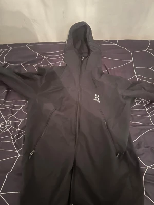 Svart vindjacka från Haglöfs - Snygg svart vindjacka från Haglöfs med huva och hel dragkedja framtill. Jackan har två stora fickor med dragkedja och diskret logga på bröstet. Tillverkad i ett lätt och slitstarkt material som passar perfekt för blåsiga dagar. Storlek m men passar perfa i s med.