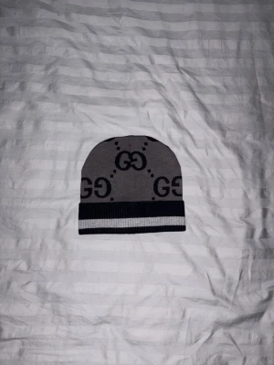 GG-mössa  - En grå och svart gg-mönstrad mössa från gucci. Hatten har ett stilrent och snyggt logomönster, och är designad med randig kant. Perfekt för dig som vill ha något trendigt och klassiskt. Mössan är diskret men ändå lyxig.