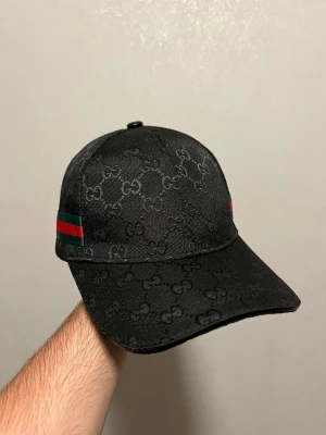 Svart Gucci keps med GG-mönster - Säljer en svart keps från Gucci med det klassiska GG-monogrammet över hela kepsen. På sidan finns Guccis ikoniska gröna och röda ränder. Kepsen är tillverkad i bomull och har justerbar rem baktill. Perfekt för dig som vill ha en stilren och lyxig accessoar.